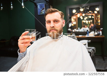Close up photo of young client holding glass of whiskey. Male customer relaxing while getting new haircut. Professional hairdresser in fancy outfit cutting client's hair. 122507556