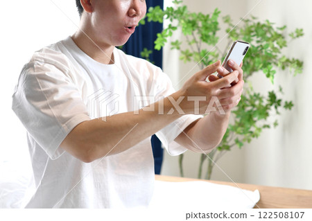 驚訝的亞洲男人坐在床上,拿著智慧型手機 驚訝的亞洲男人坐在床上,拿著智慧型手機 122508107
