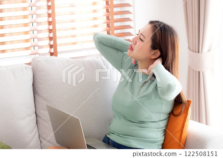 Asian woman sitting on sofa with neck pain while using laptop computer in living room. 122508151