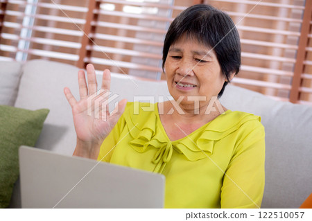 Senior asian woman waving during a video call on laptop computer sitting on sofa in living room. 122510057