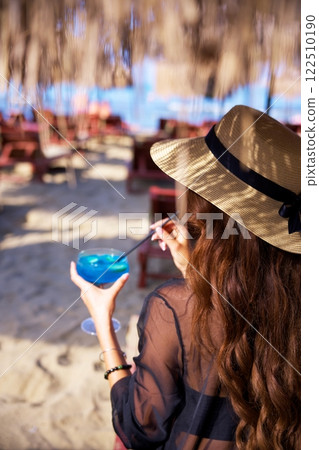 Woman in sun hat enjoying tropical cocktail at beachside bar with ocean view  122510190