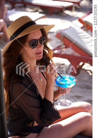 Stylish woman in hat enjoying blue cocktail at beach  122510195