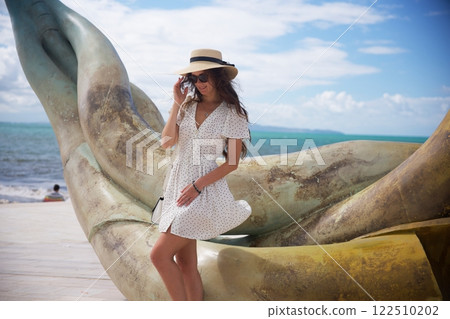 Smiling woman in sun hat posing by artistic seaside sculpture  122510202