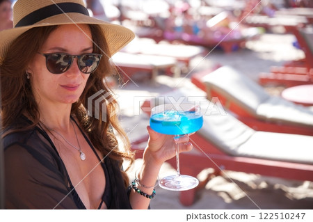 Chic woman holding a cold blue cocktail on sunny beach Chic woman holding a cold blue cocktail on sunny beach 122510247