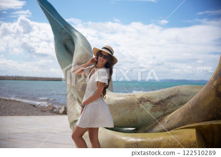 Smiling woman in sun hat posing by seaside sculpture Smiling woman in sun hat posing by seaside sculpture 122510289