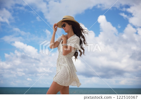 Elegant woman in sun hat adjusting hat in ocean breeze Elegant woman in sun hat adjusting hat in ocean breeze 122510367