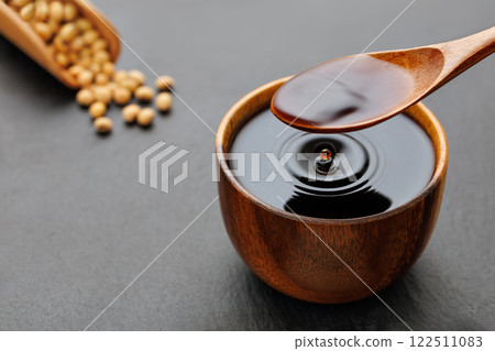 soy sauce pouring from spoon in bowl with splash and drop and dry soybeans on black stone background 122511083