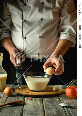 In a warm, rustic kitchen, a chef skillfully prepares a mixture by cracking an egg into a bowl of milk. The wooden surface showcases ingredients, enhancing the culinary atmosphere In a warm, rustic kitchen, a chef skillfully prepares a mixture by cracking an egg into a bowl of milk. The wooden surface showcases ingredients, enhancing the culinary atmosphere 122516979