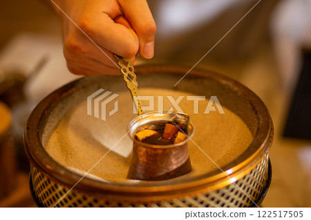 A barista's hand skillfully crafts coffee on sand using a traditional copper Turkish cezve 122517505
