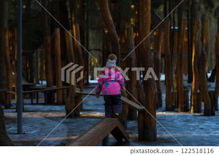 Young child in colorful winter clothing carefully balances on a wooden beam surrounded by tall trees in a serene forest playground, creating a sense of adventure and exploration. Young child in colorful winter clothing carefully balances on a wooden beam surrounded by tall trees in a serene forest playground, creating a sense of adventure and exploration. 122518116