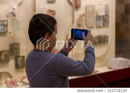 The young student explores the museum exhibition and takes pictures of artifacts with a smartphone, enjoying cultural and educational experience. A teenager at the museum on excursions. The young student explores the museum exhibition and takes pictures of artifacts with a smartphone, enjoying cultural and educational experience. A teenager at the museum on excursions. 122518120