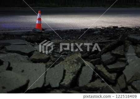 A traffic cone placed near broken asphalt on a damaged road at night. The illuminated road contrasts with the dark debris, emphasizing urban roadwork and construction hazards. 122518409