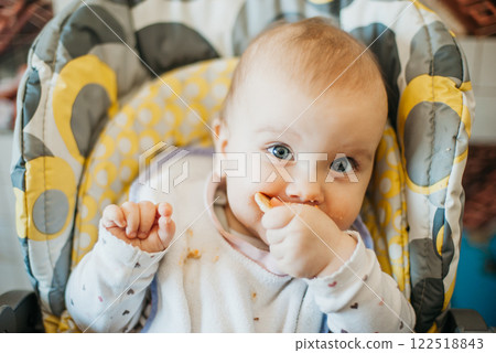 Cute Baby with Messy Face Eating Solid Food During Mealtime. Baby sitting in a high chair with food on their face, holding a piece of food. Early self-feeding and baby nutrition concept Cute Baby with Messy Face Eating Solid Food During Mealtime. Baby sitting in a high chair with food on their face, holding a piece of food. Early self-feeding and baby nutrition concept 122518843