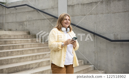 Happy young woman with mobile standing on steps in city. Holidays, technology and tourism concept. Female tourist searching direction on smart phone while traveling abroad in summer 122518902