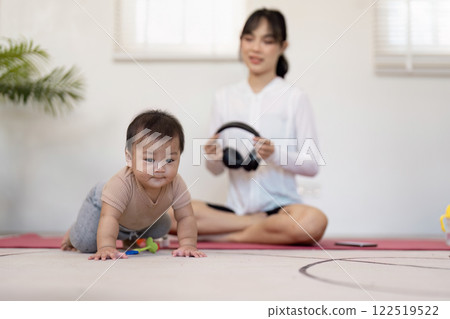 Multitasking mother supervising her crawling baby while preparing for meditation with headphones at home 122519522