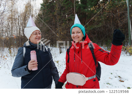 Two friends stroll through a serene, snow-covered forest, donning playful party hats. The atmosphere is cheerful 122519566