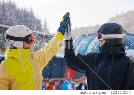 Two skiers in colorful ski gear smiling and slapping each other on the palm and having fun with snow. Ski resort concept, 122521004