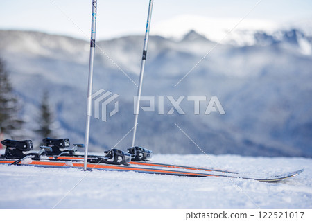 A pair of skis and poles on a snowy mountain slope with a blurred mountain range in the background. 122521017