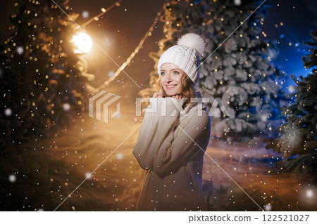 Woman in white winter hat and sweater warming her hands in snowy winter forest with blurred Christmas lights in background. Woman in white winter hat and sweater warming her hands in snowy winter forest with blurred Christmas lights in background. 122521027
