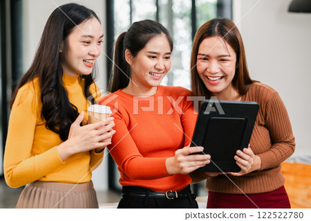 Three young women smiling and looking at a tablet together in a bright, modern office setting. Three young women smiling and looking at a tablet together in a bright, modern office setting. 122522780