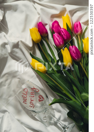Wine glass with words HELLO SPRING Fresh bouquet of colorful tulip flowers on bed. Gift present idea for holiday birthday 8 March Mother's Day Valentine's day Women's Day 122530397
