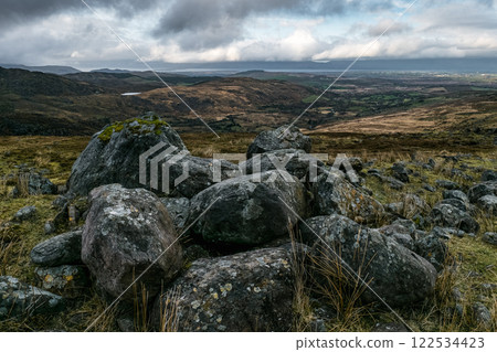 Grey boulders, some moss-covered, are scattered across a hillside. The background shows a vast, rolling landscape under a cloudy sky. Grey boulders, some moss-covered, are scattered across a hillside. The background shows a vast, rolling landscape under a cloudy sky. 122534423