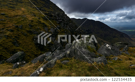 Rough terrain with rocks and vegetation, overcast sky above the mountains. 122534435