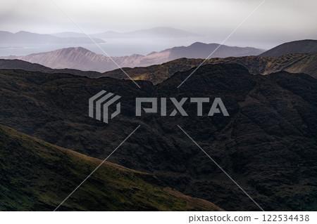 Layers of dark hills create a dramatic mountain landscape, shrouded in mist as the sun sets, revealing a distant coastline against a moody sky. Layers of dark hills create a dramatic mountain landscape, shrouded in mist as the sun sets, revealing a distant coastline against a moody sky. 122534438
