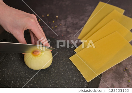 Woman's hand cutting onion on the black kitchen cutting board. Lasagne pasta sheets. Black background. Homemade lasagna cooking preparation 122535163