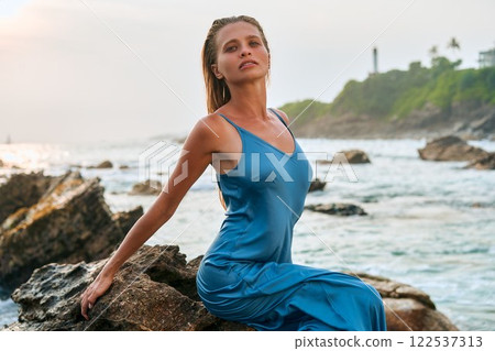 Model woman in wet dress sits on rock by tropical sea with waves. Ocean storm, exotic island background. Luxury fashion, beauty advertisement. Romantic, dreamy vibes. Model woman in wet dress sits on rock by tropical sea with waves. Ocean storm, exotic island background. Luxury fashion, beauty advertisement. Romantic, dreamy vibes. 122537313