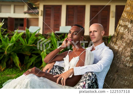 Ambiguous gender fluid person in bride dress with LGBTQ groom in bold suit at tropical villa wedding. Celebrate diversity, love, inclusion. Beachside ceremony, modern fashion, joyful moment. 122537318