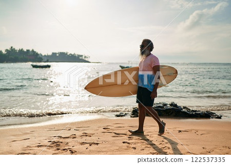 Active senior ethnic man walks on sunny beach with surfboard. Elderly surfer enjoys ocean waves in vibrant lifestyle. Retirement adventure with surf culture. Promoting healthy aging in coastal scene. 122537335