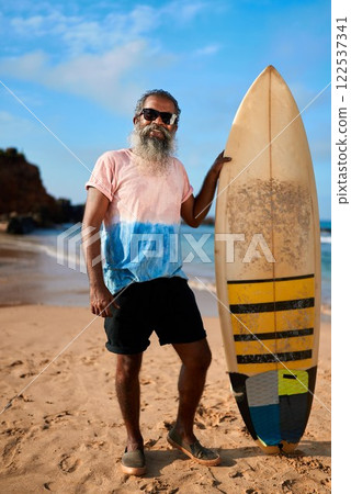 Senior man with grey beard stands on sandy beach holding surfboard. Elderly surfer enjoys sunny day by ocean. Retirement active lifestyle and wellness vibes. Happy golden years at seaside. 122537341