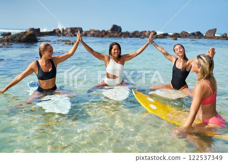 Four women friends on surfboards hold hands sitting in clear water. Smiling, bonding, and enjoying sunny day. Surf camp vibes with ocean backdrop. Strong female friendship and water sports connection. 122537349