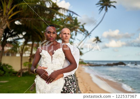 Ambiguous gender fluid person in white wedding dress with lgbtq partner in bold suit on beach. Tropical villa, palms, ocean. Gay wedding event, honeymoon travel tour theme. 122537367