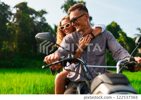 Couple rides motorbike through green field. Romantic adventure, exploring tropical countryside. Perfect for honeymoon, travel, Valentine Day. Man drives scooter, woman hugs. Couple rides motorbike through green field. Romantic adventure, exploring tropical countryside. Perfect for honeymoon, travel, Valentine Day. Man drives scooter, woman hugs. 122537368