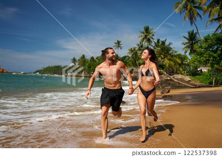 Happy couple runs on tropical beach, hand in hand. Healthy young man, woman in swimwear jog on sandy shore, palm trees in background. Fitness run, sunny travel destination. Active vacation, wellness. 122537398