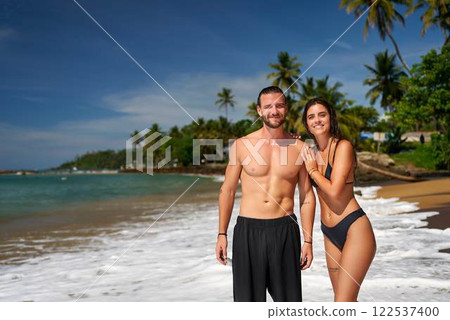 Fit couple stands on tropical beach, man shirtless with toned abs, woman in bikini, both smiling. Active pair enjoys sunny seaside, health, travel lifestyle. Vacation at exotic destination by sea. 122537400