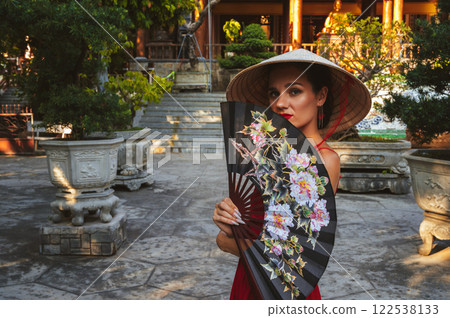 Portrait of a female tourist with a fan in a Vietnamese traditional non la hat at pagoda on a trip to Vietnam in Asia Portrait of a female tourist with a fan in a Vietnamese traditional non la hat at pagoda on a trip to Vietnam in Asia 122538133