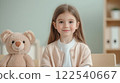 young girl with long hair sits at table with teddy bear, smiling warmly in cozy classroom setting 122540667