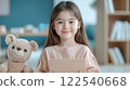 smiling young girl holding card sits next to teddy bear in cozy, well lit room 122540668