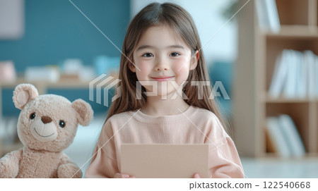 smiling young girl holding card sits next to teddy bear in cozy, well lit room 122540668