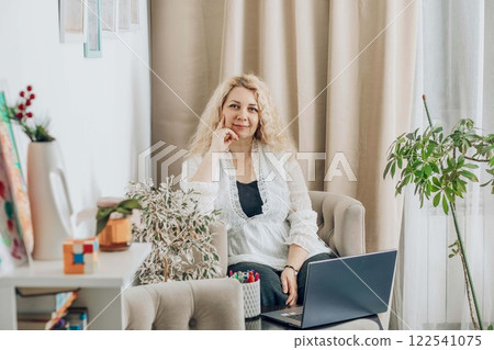 woman with laptop in cozy home office on beige armchair, with plants and in white blouse with curly blonde hair, concept of remote work, business, lifestyle woman with laptop in cozy home office on beige armchair, with plants and in white blouse with curly blonde hair, concept of remote work, business, lifestyle 122541075