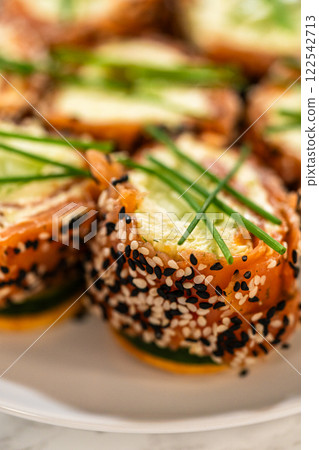 A close-up shot of smoked salmon jumbo cream cheese rolls artfully arranged on a white plate. The rolls are wrapped in vibrant smoked salmon, sprinkled with black and white sesame seeds, and topped 122542713