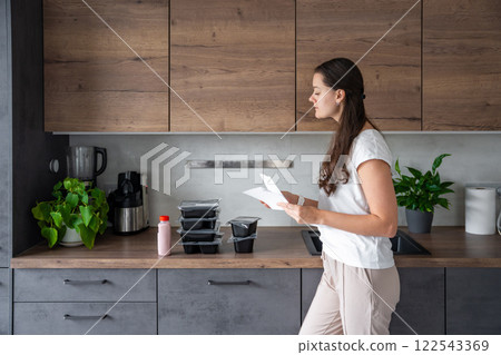 Young woman with healthy diet plan for weight loss, delivery of daily ready meal menu. Woman using meal tracker app on phone. Balanced portion with dish. Pre-cooking. Young woman with healthy diet plan for weight loss, delivery of daily ready meal menu. Woman using meal tracker app on phone. Balanced portion with dish. Pre-cooking. 122543369