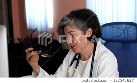 Caucasian doctor with short gray hair, lifting glasses slightly, gentle smile showing attention and focus, composed expression, thinking deeply during medical discussion. 122543627