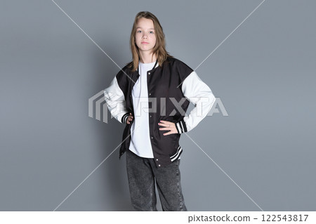 A young girl confidently carries her varsity jacket on one shoulder while facing sideways. The natural expression reflects a calm and relaxed attitude. A young girl confidently carries her varsity jacket on one shoulder while facing sideways. The natural expression reflects a calm and relaxed attitude. 122543817