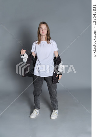 A teenage girl strikes a casual pose, wearing jeans and a varsity jacket over a white shirt. Her outfit represents youthful confidence and the freedom of self-expression in everyday wear. A teenage girl strikes a casual pose, wearing jeans and a varsity jacket over a white shirt. Her outfit represents youthful confidence and the freedom of self-expression in everyday wear. 122543831