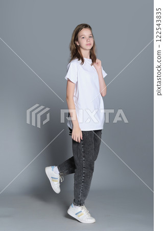 A teenage girl stands confidently with one leg raised slightly, wearing a white t-shirt, jeans, and a jacket over her shoulder. Her relaxed posture reflects a casual and youthful sense of style. 122543835