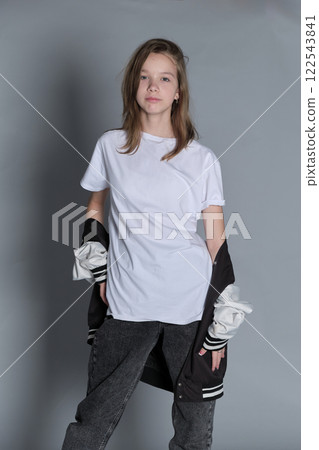 A teenage girl stands confidently in a white t-shirt and jeans, holding a jacket loosely at her side. Her relaxed expression and casual pose reflect a laid-back attitude and effortless style. A teenage girl stands confidently in a white t-shirt and jeans, holding a jacket loosely at her side. Her relaxed expression and casual pose reflect a laid-back attitude and effortless style. 122543841